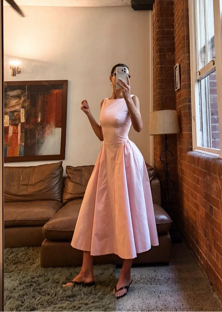 Woman in a pink dress taking a mirror selfie in a room with a large window and brick wall.